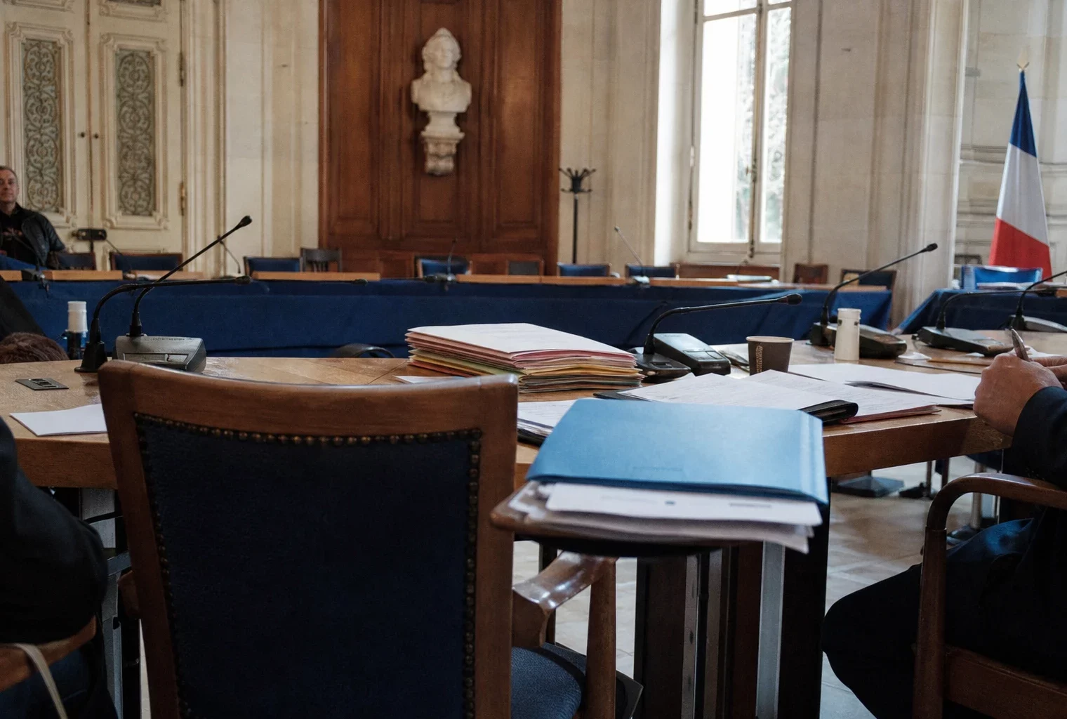 Salle du conseil municipal à Nice, avec chaise vide, micros et dossiers sur une longue table éclairée par la lumière du jour.