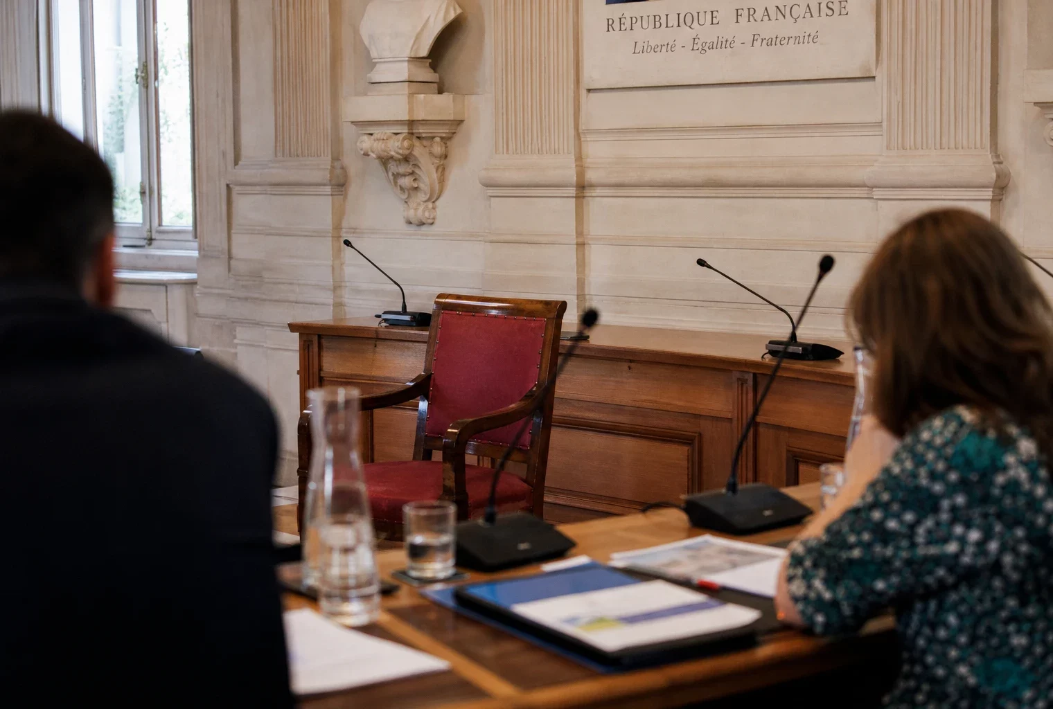 Salle municipale claire avec une chaise vide, des micros et des dossiers flous autour d’une table de conseil local.