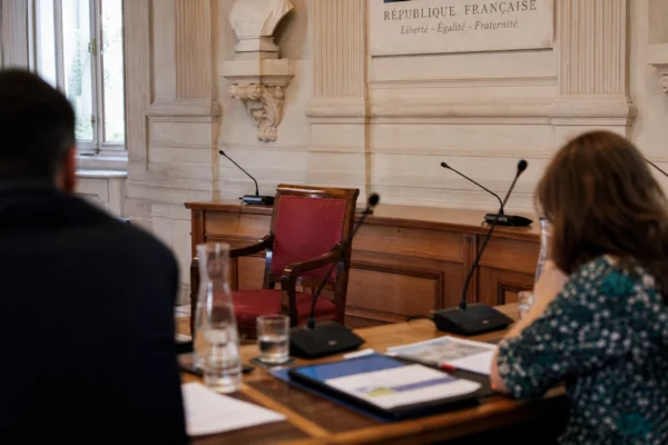 Salle municipale claire avec une chaise vide, des micros et des dossiers flous autour d’une table de conseil local.