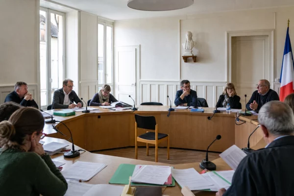 Salle municipale lumineuse avec chaise vide, micros et dossiers flous, en ambiance institutionnelle discrète.