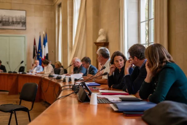 Salle municipale lumineuse avec micros, dossiers et une chaise vide lors d’une réunion locale