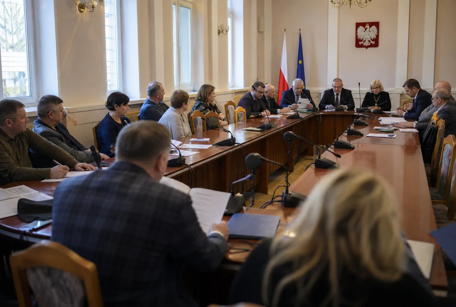 Salle municipale polonaise avec chaise vide, micros et dossiers flous, en lumière naturelle, ambiance de réunion politique locale.