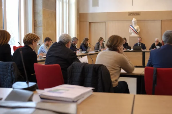 Salle municipale française lumineuse avec micros, dossiers flous et chaise vide lors d’une réunion locale.