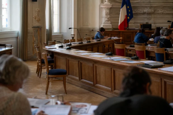 Salle municipale lumineuse avec micros, dossiers flous et chaise vide avant une audience liée à Sébastien Delogu