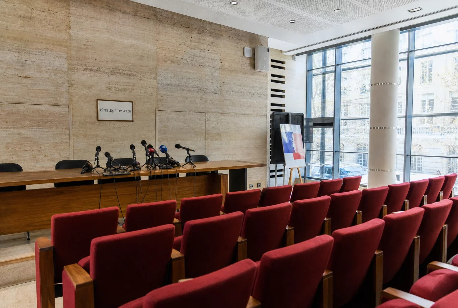 Salle de presse institutionnelle vide avec pupitre, micros et rangées de sièges rouges, éclairage naturel clair.