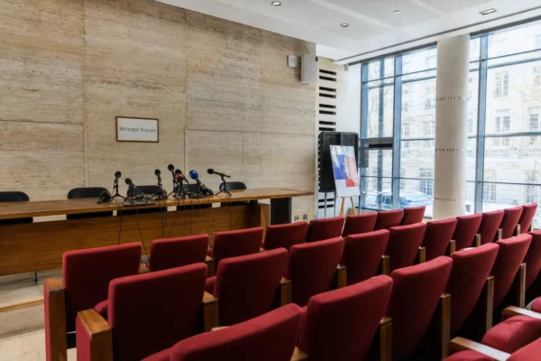 Salle de presse institutionnelle vide avec pupitre, micros et rangées de sièges rouges, éclairage naturel clair.