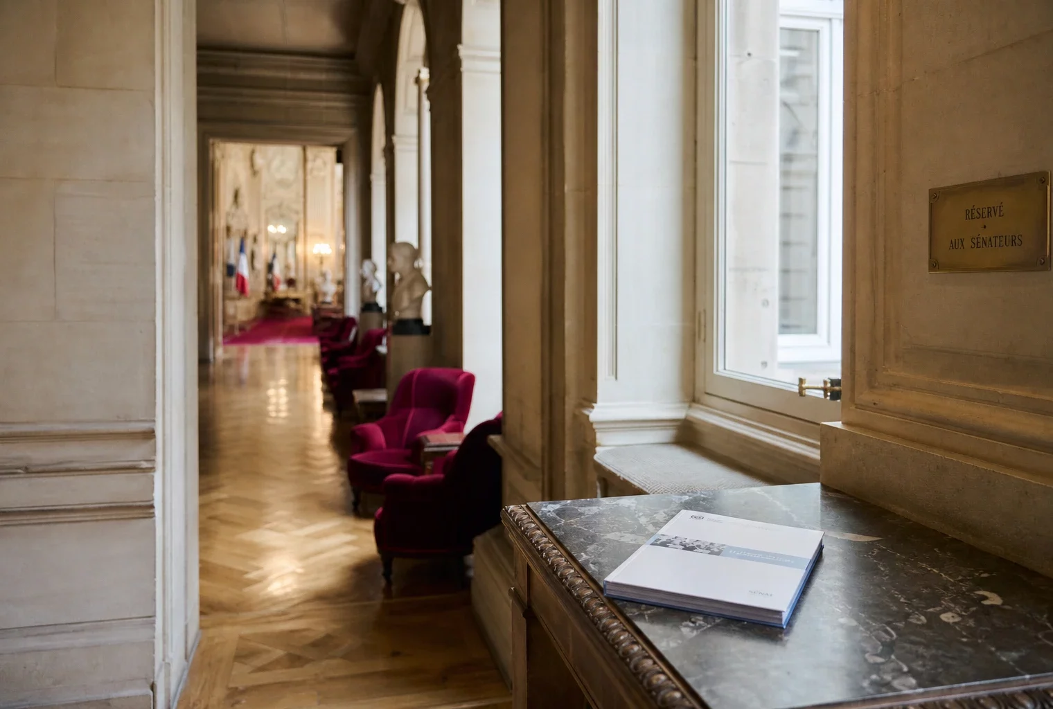 Couloir du Sénat vide avec marbre clair, sièges rouges et dossier législatif posé sur une table.