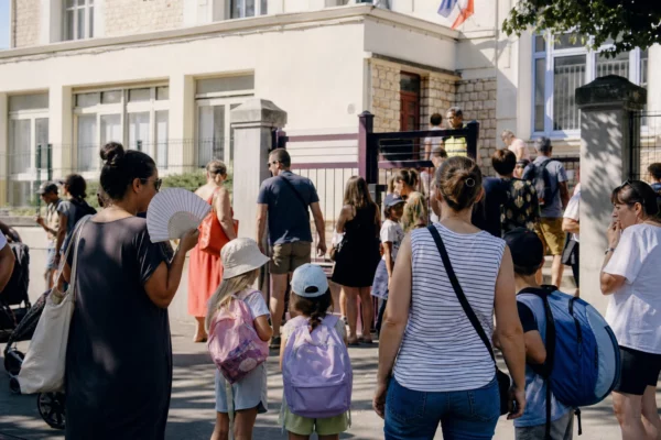 Parents anonymes à la sortie d’une école publique française par forte chaleur, sous une lumière de matin claire.