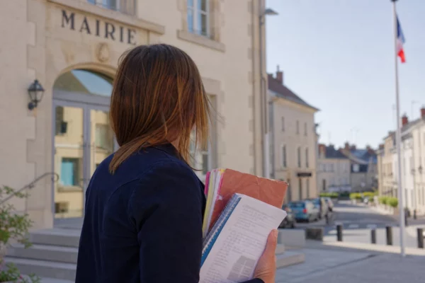Collaborateur municipal anonyme devant une mairie française, dossier en main, sur fond de rue calme