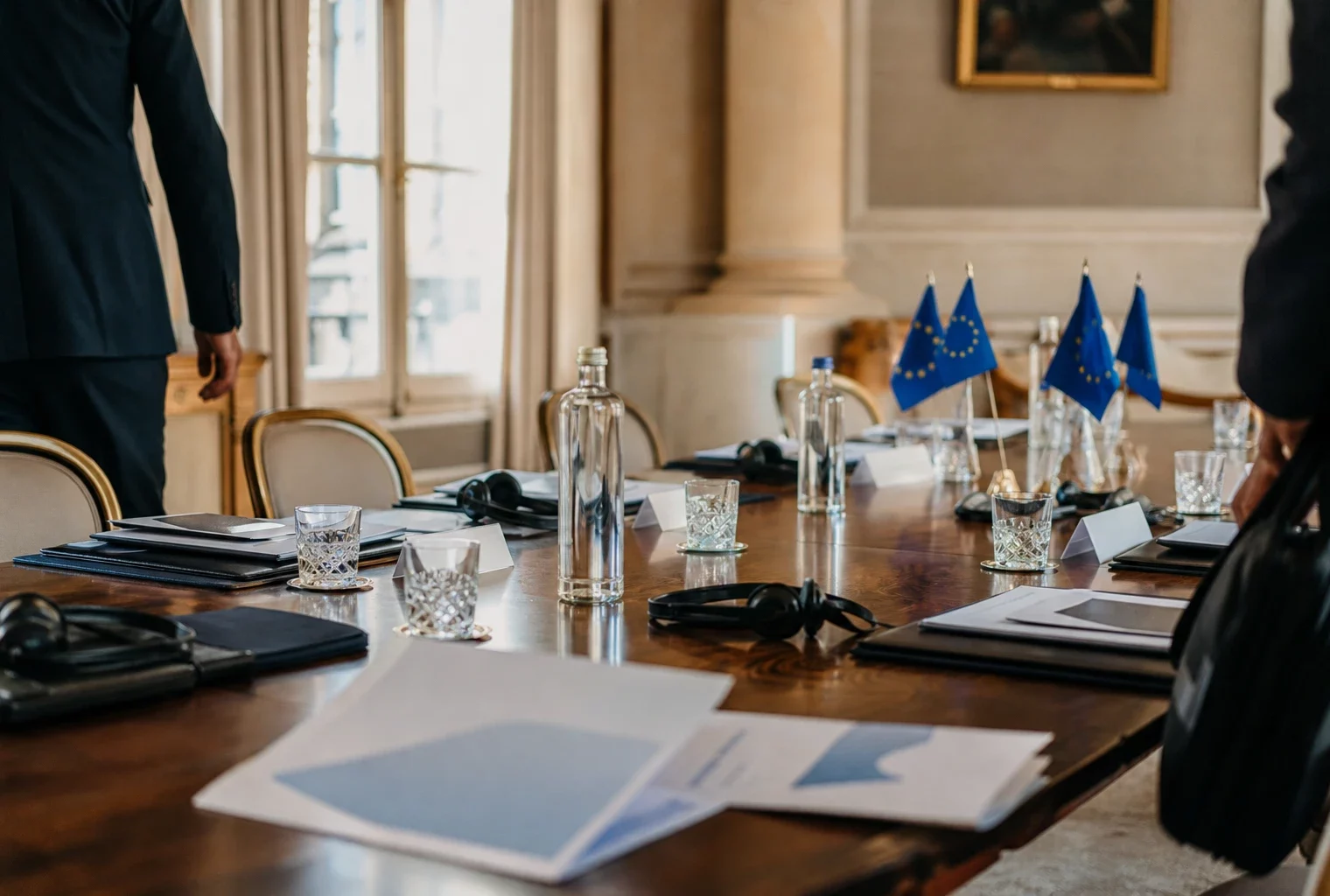Table de négociation institutionnelle avec dossiers, écouteurs, verres d’eau et drapeaux européens discrets.