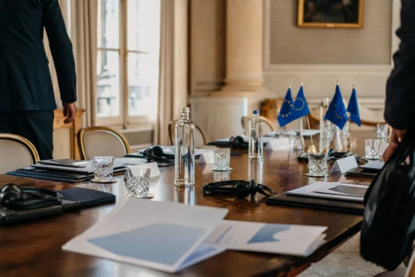 Table de négociation institutionnelle avec dossiers, écouteurs, verres d’eau et drapeaux européens discrets.