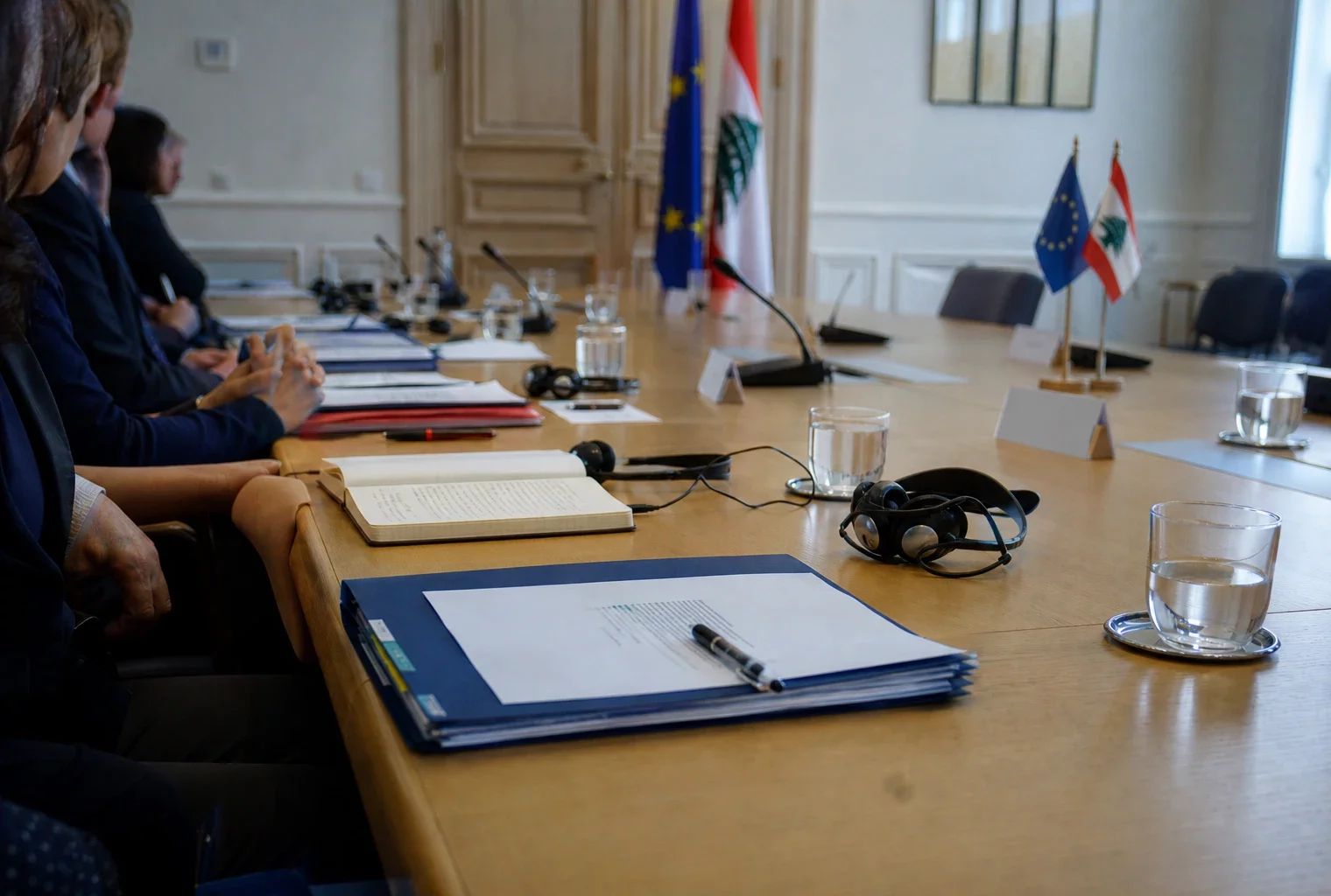 Table de négociation européenne avec dossiers, écouteurs et drapeaux miniatures, dans une salle diplomatique lumineuse.