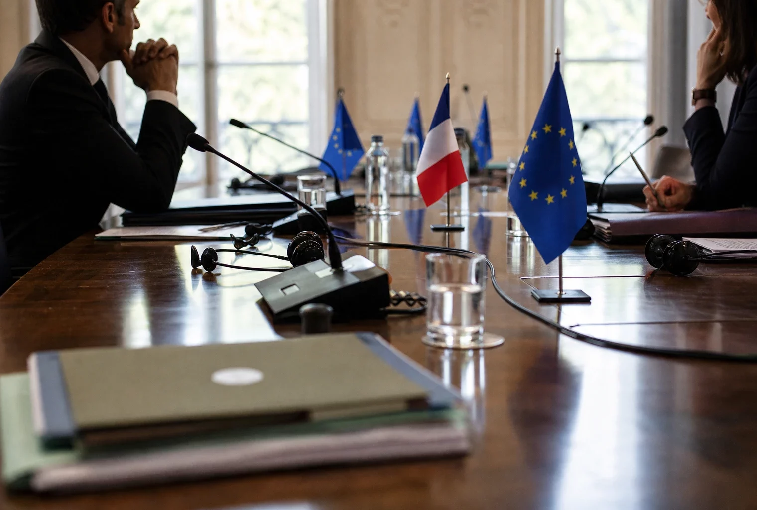 Table diplomatique européenne avec dossiers, écouteurs, verres d'eau et mini-drapeaux, silhouettes anonymes en réunion