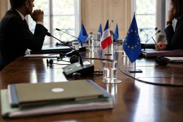 Table diplomatique européenne avec dossiers, écouteurs, verres d'eau et mini-drapeaux, silhouettes anonymes en réunion