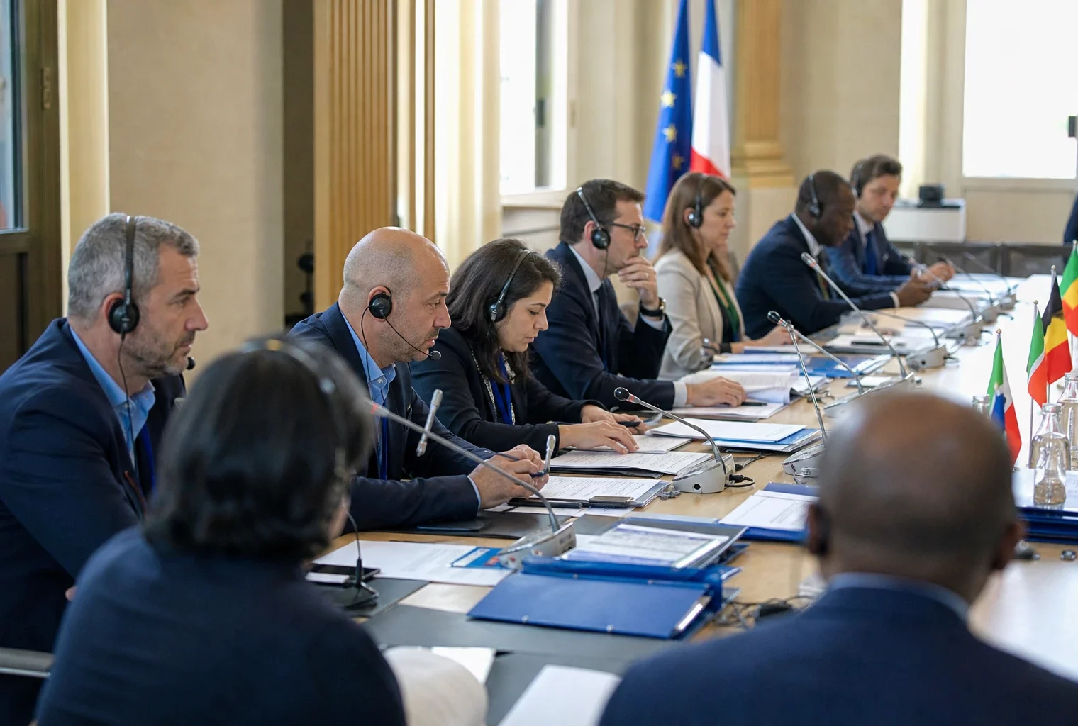 Réunion internationale autour d’une table de négociation, avec micros, dossiers et silhouettes anonymes sous une lumière claire.