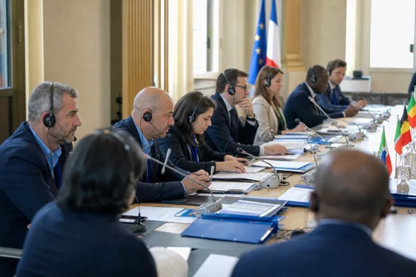 Réunion internationale autour d’une table de négociation, avec micros, dossiers et silhouettes anonymes sous une lumière claire.
