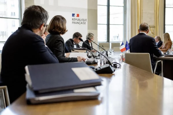 Table de négociation diplomatique avec dossiers, micros et silhouettes anonymes dans un cadre institutionnel français.