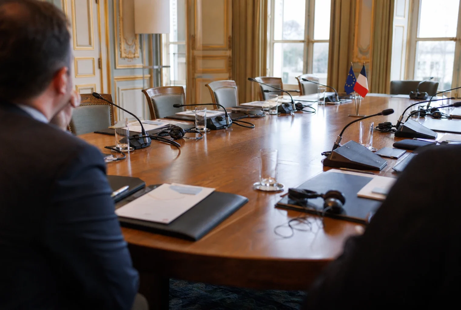 Table de négociation diplomatique à Paris avec dossiers, micros et silhouettes anonymes dans une lumière naturelle.