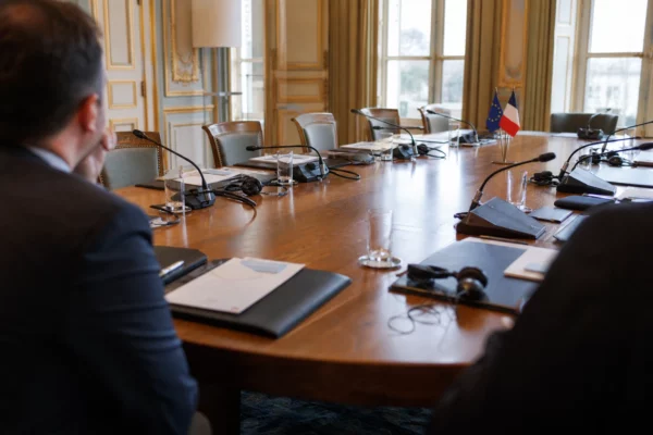 Table de négociation diplomatique à Paris avec dossiers, micros et silhouettes anonymes dans une lumière naturelle.