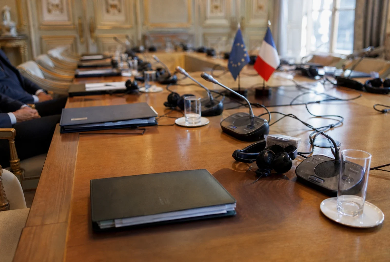 Table de négociation avec dossiers, micros et silhouettes anonymes dans une salle institutionnelle lumineuse