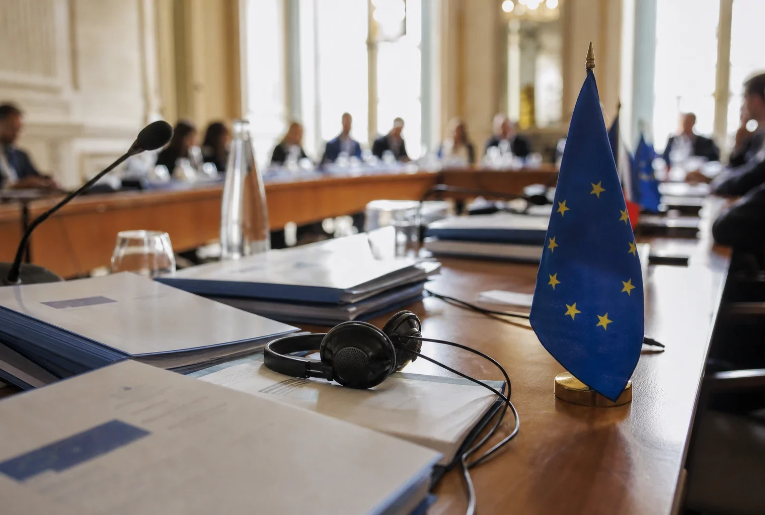 Table de négociation européenne avec dossiers, écouteurs et petits drapeaux de l’UE dans une salle institutionnelle lumineuse.