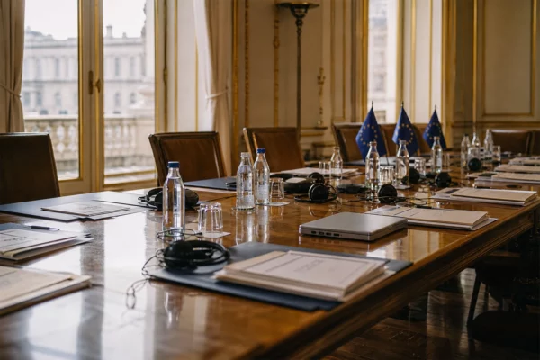 Table de négociation européenne avec dossiers, casques, verres d’eau et petits drapeaux de table discrets.