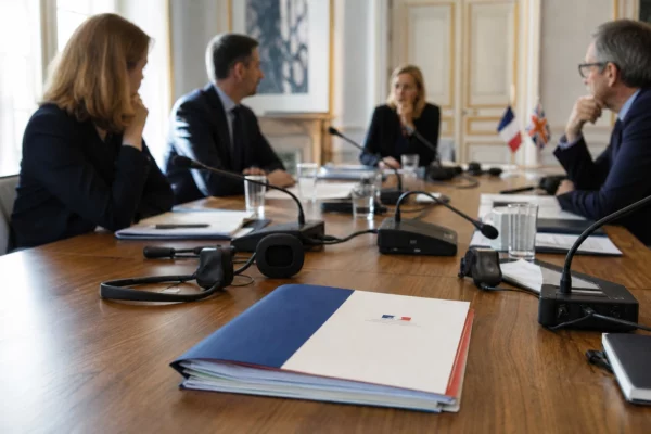 Table de négociation diplomatique avec dossiers, micros et petits drapeaux français et britannique, dans une salle institutionnelle claire.