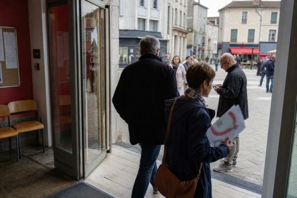 Des personnes anonymes entrent dans un centre culturel municipal en ville moyenne française, avec affiches floues et lumière naturelle.
