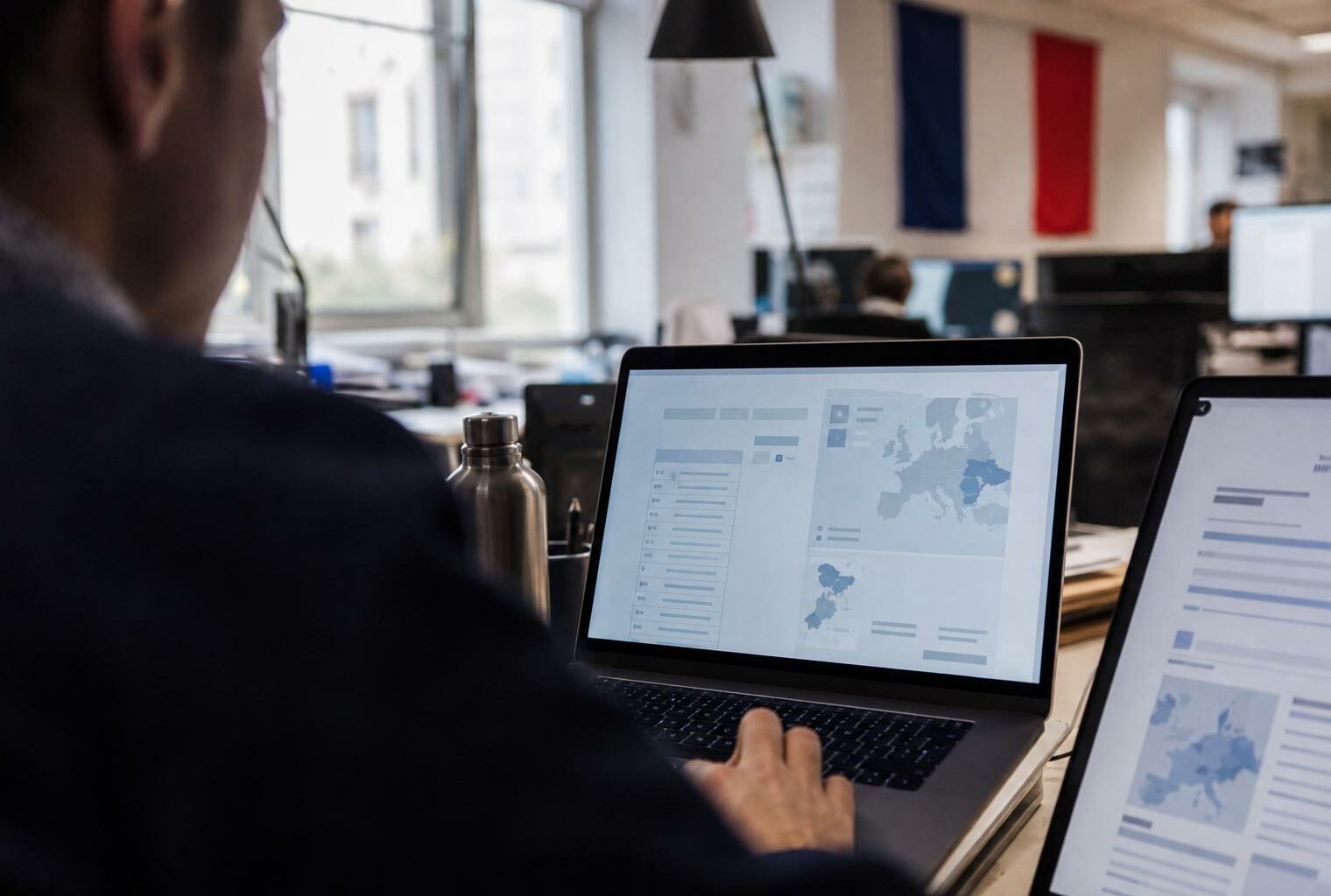 Journaliste vu de dos devant un ordinateur affichant une interface européenne abstraite, dans une rédaction lumineuse