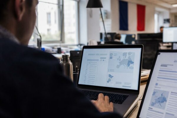 Journaliste vu de dos devant un ordinateur affichant une interface européenne abstraite, dans une rédaction lumineuse