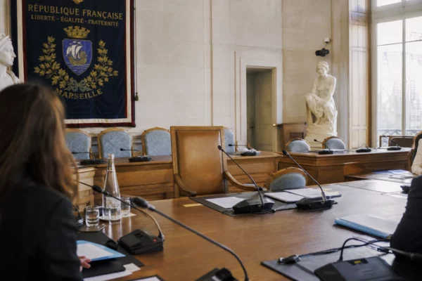 Salle municipale française avec chaise vide, micros et dossiers, dans une ambiance claire de briefing diplomatique.