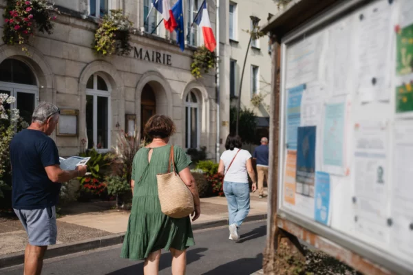 Des habitants anonymes passent devant une mairie française par une belle lumière de jour, avec un panneau d’affichage flou.