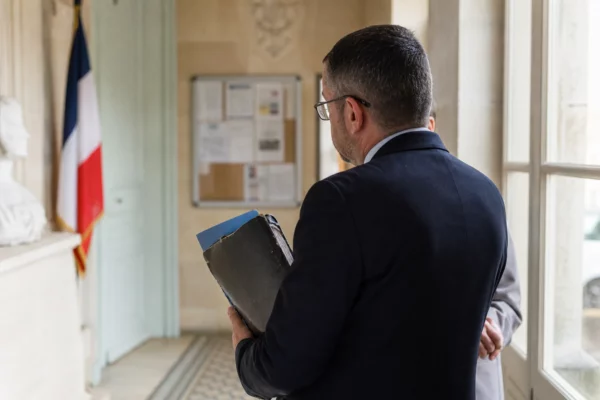 Un élu local vu de dos dans une mairie, carnet en main, sur fond de dossier et de lumière naturelle.