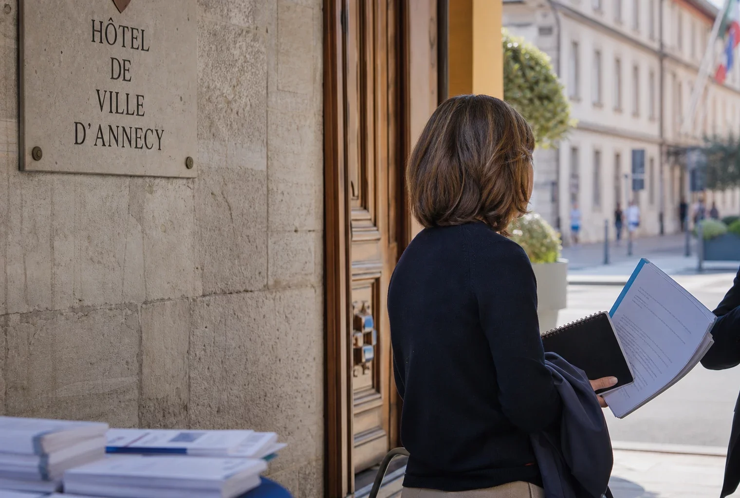 Un élu local de dos échange avec un collègue devant la mairie d’Annecy, dossier et carnet en main.