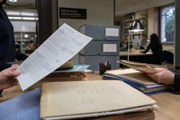 Dans des archives publiques françaises, des mains anonymes consultent des dossiers administratifs sous une lampe claire.