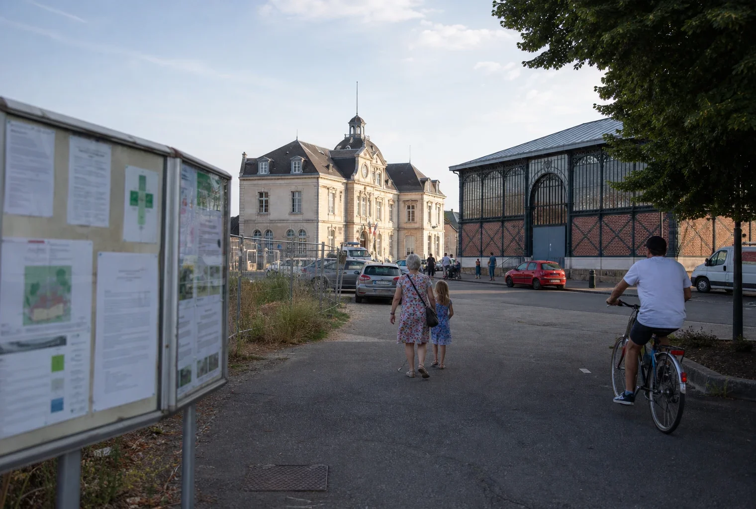 Scène de ville moyenne française avec marché, mairie locale et terrain en reconversion sous une lumière claire.