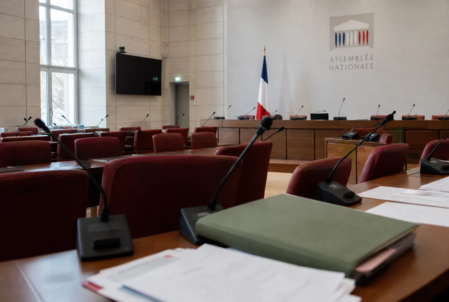 Salle de commission vide à l’Assemblée nationale avec micros et dossier agricole fermé sous la lumière du jour.