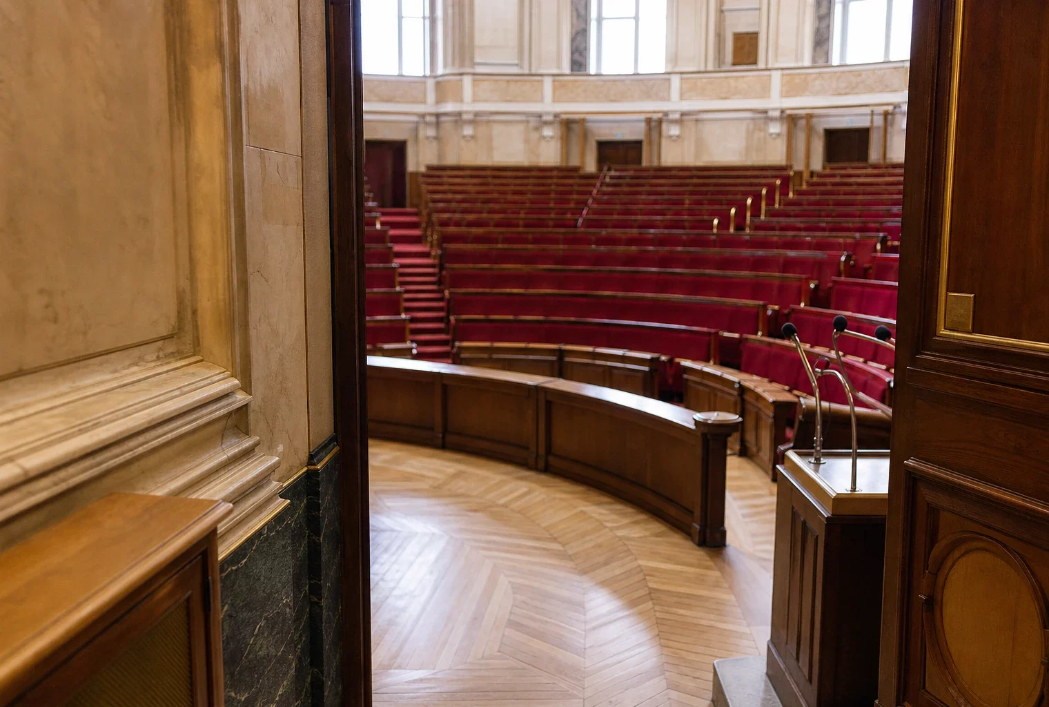 Couloir de l’Assemblée nationale avec vue oblique vers l’hémicycle, sièges rouges et lumière naturelle claire.