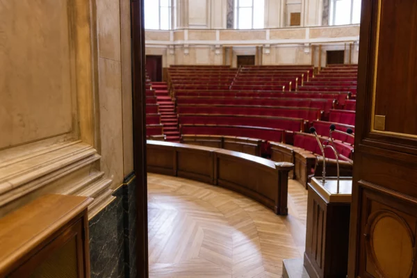 Couloir de l’Assemblée nationale avec vue oblique vers l’hémicycle, sièges rouges et lumière naturelle claire.