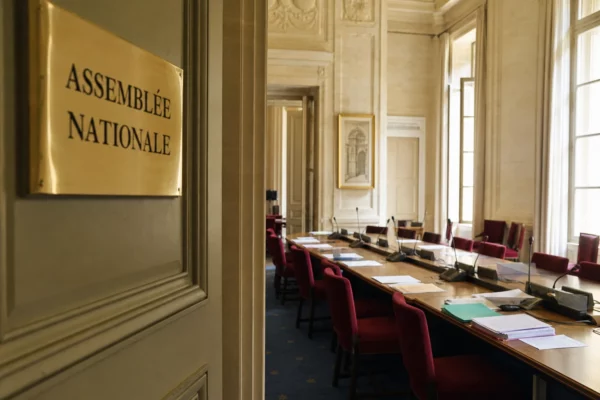 Salle de commission vide à l’Assemblée nationale, avec sièges rouges et micros sous une lumière naturelle claire.