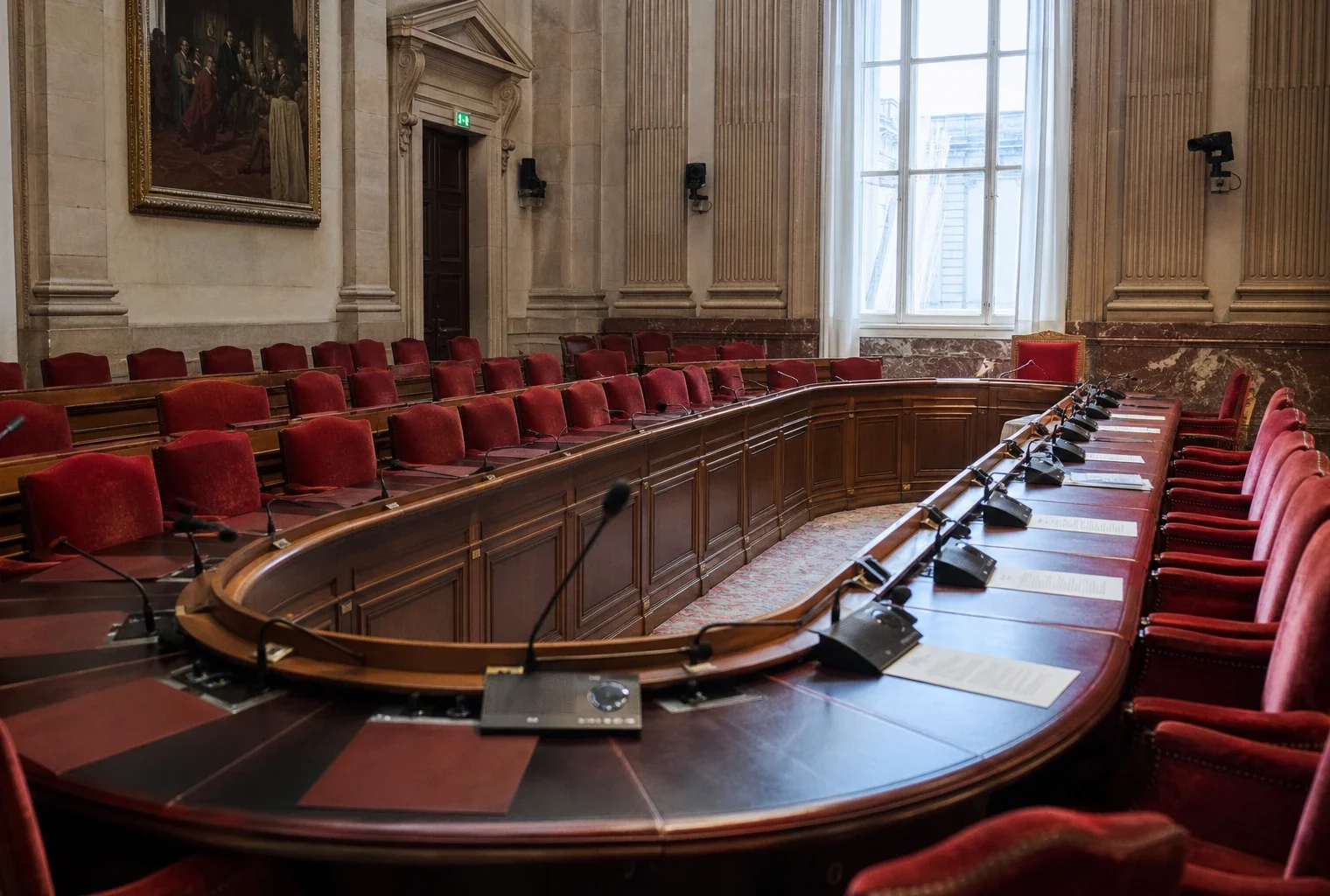 Salle de commission de l’Assemblée nationale vide, avec micros et dossiers sur la table, vue oblique en lumière naturelle.