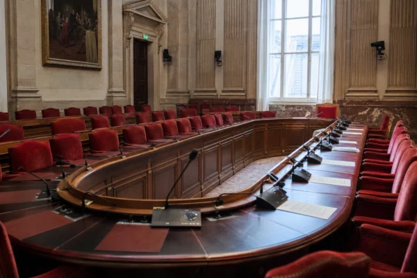 Salle de commission de l’Assemblée nationale vide, avec micros et dossiers sur la table, vue oblique en lumière naturelle.