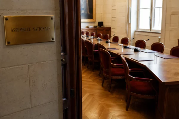Salle institutionnelle vide à l’Assemblée nationale, avec tables, micros et dossiers dans une lumière naturelle claire.