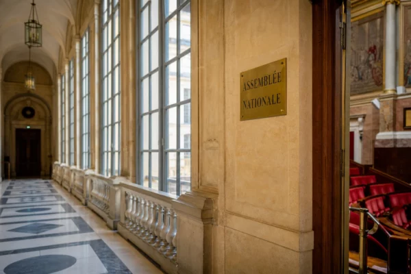 Couloir vide de l’Assemblée nationale en lumière naturelle, avec les sièges rouges du Palais-Bourbon en arrière-plan.