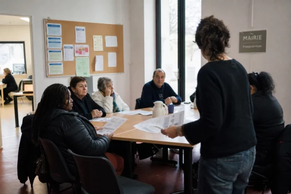 Réunion locale dans une association de quartier, avec des habitants anonymes autour de documents flous sur le vote.