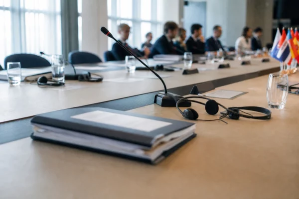 Table de négociation internationale avec dossiers fermés, micros et silhouettes anonymes dans une salle lumineuse.