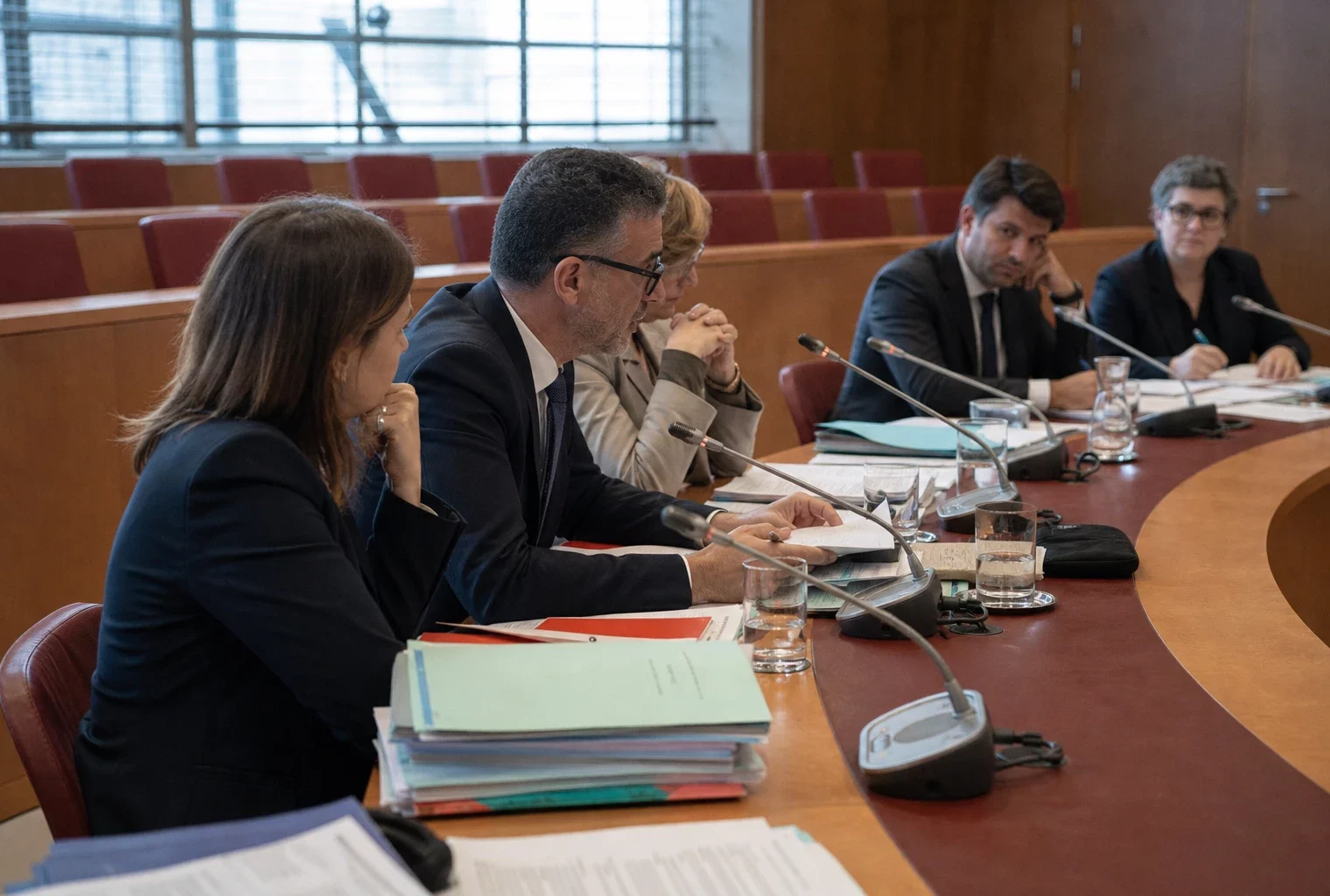 Réunion de commission des finances à l’Assemblée, avec micros, dossiers ouverts et silhouettes anonymes sous une lumière claire.