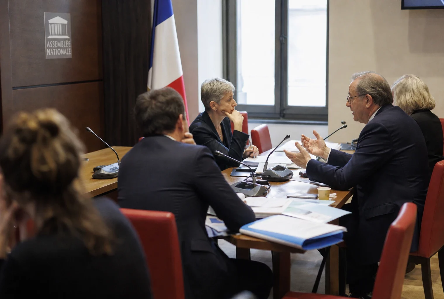 Réunion de commission à l’Assemblée avec micros, dossiers ouverts et silhouettes anonymes en lumière naturelle.