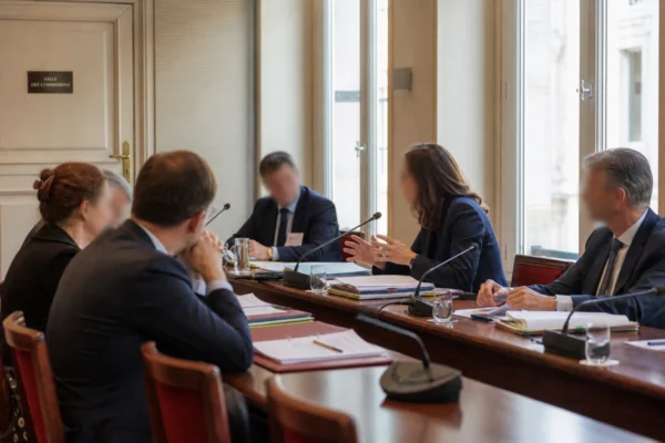 Réunion de commission parlementaire avec micros, dossiers ouverts et silhouettes anonymes dans une salle lumineuse.