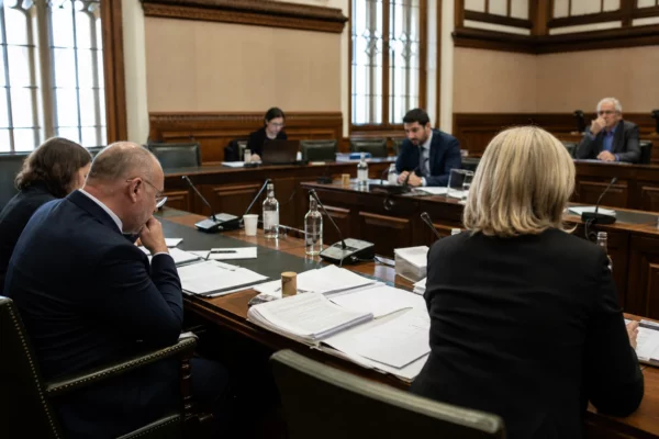 Réunion de commission parlementaire avec micros, dossiers ouverts et silhouettes anonymes dans une salle claire.